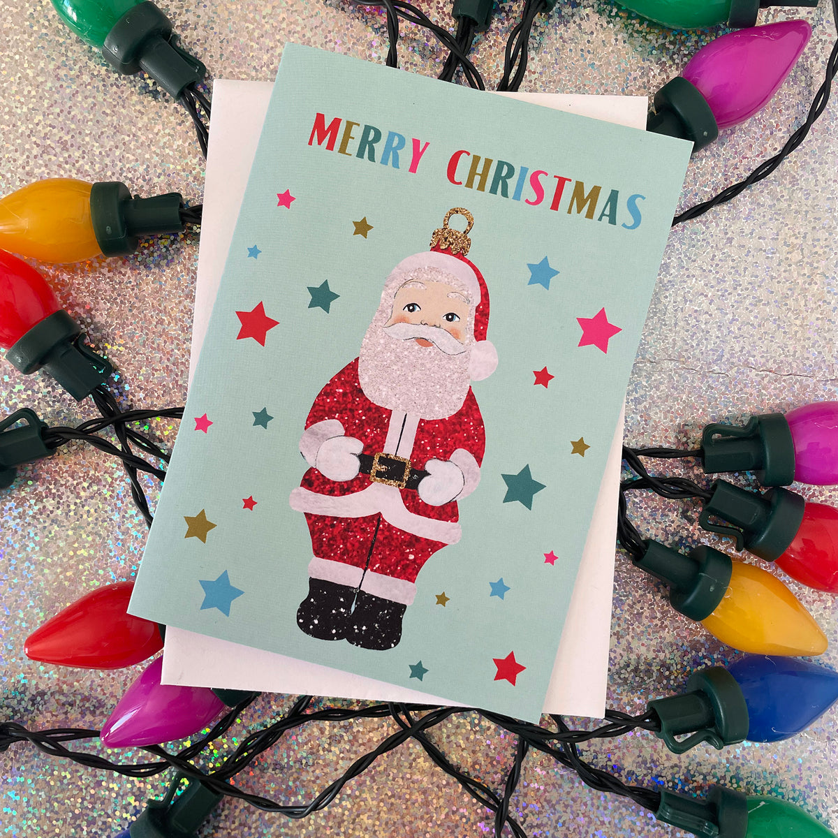 A Christmas greeting card featuring a glittery Santa Claus illustration and the words 'MERRY CHRISTMAS' in multicoloured letters, placed on top of a white envelope and surrounded by colourful Christmas lights on a sparkly silver background.