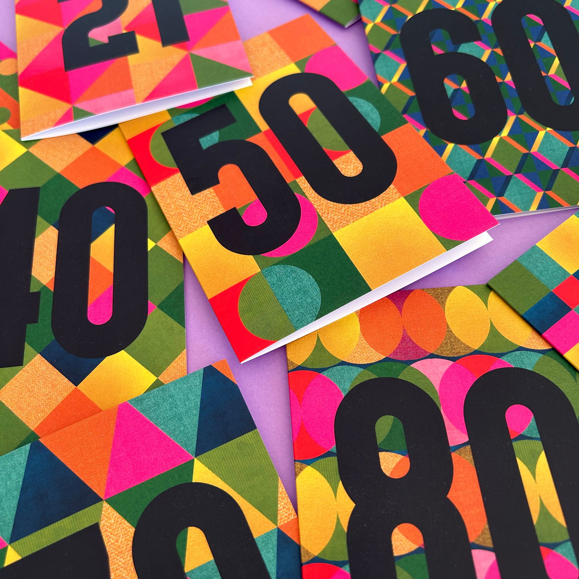 A close-up flat lay of colourful patterned cards or paper pieces featuring large black numbers such as 50, 60, 40, 80, and 21 over bright geometric and circular designs in pink, orange, yellow, green, blue, and purple.