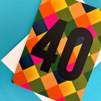 A bright greeting card with a colourful geometric pattern of overlapping coloured squares and a large black '40' printed across the front, shown on a bright blue background with a white envelope partially visible underneath.