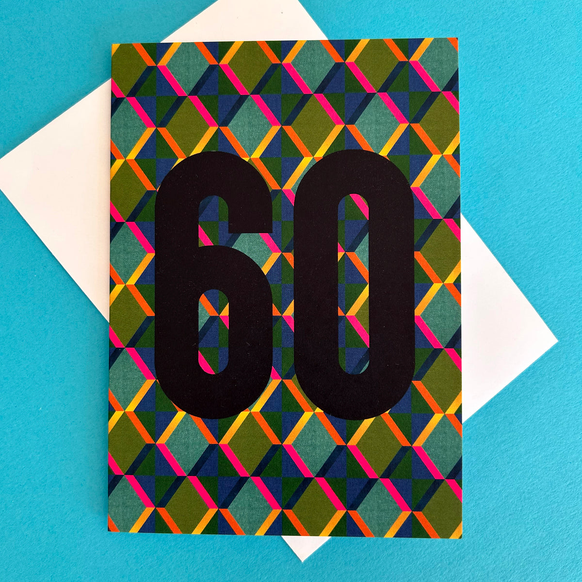 A colourful greeting card with a bold geometric pattern in green, orange, pink, yellow, and blue, featuring the number '60' in large black font. The card is displayed on top of a white envelope against a bright blue background.