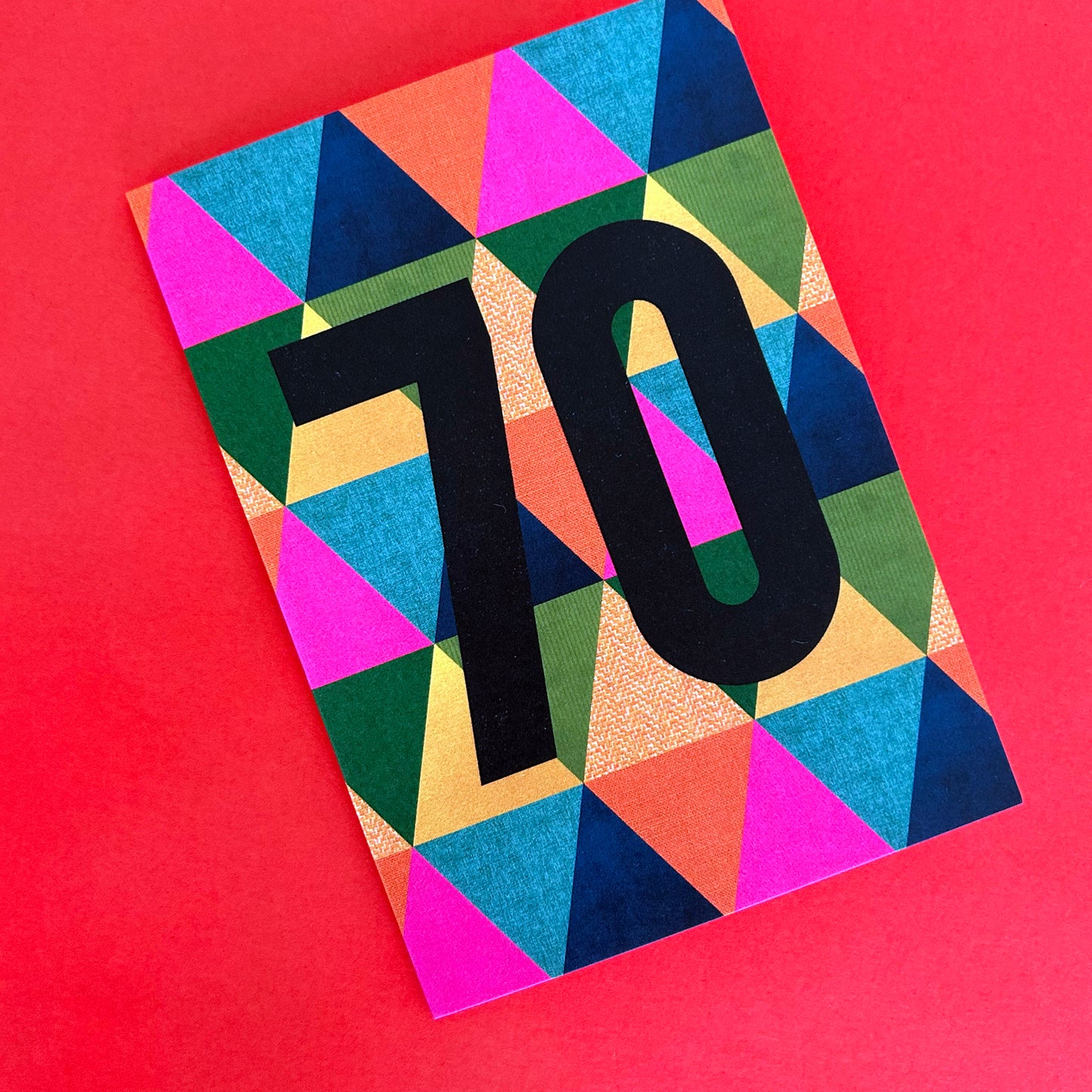 A bright greeting card featuring a bold black '70' on a vibrant, multicoloured geometric triangle pattern, set against a solid red background.