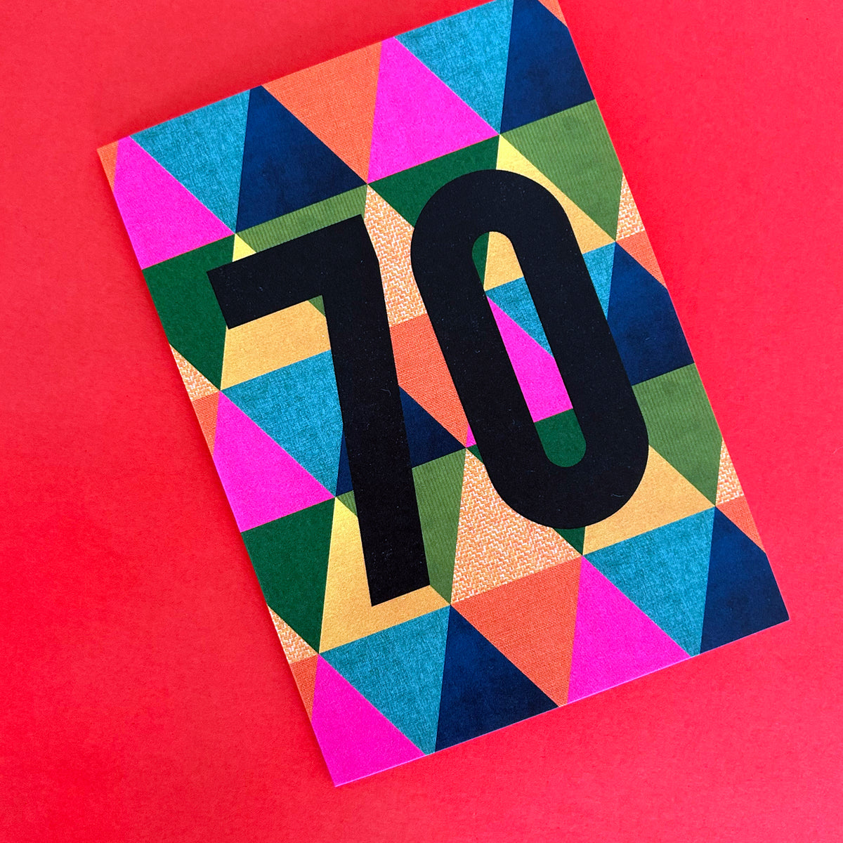 A bright greeting card featuring a bold black '70' on a vibrant, multicoloured geometric triangle pattern, set against a solid red background.