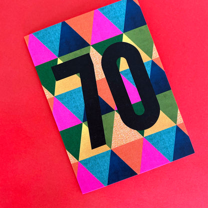 A bright greeting card featuring a bold black '70' on a vibrant, multicoloured geometric triangle pattern, set against a solid red background.
