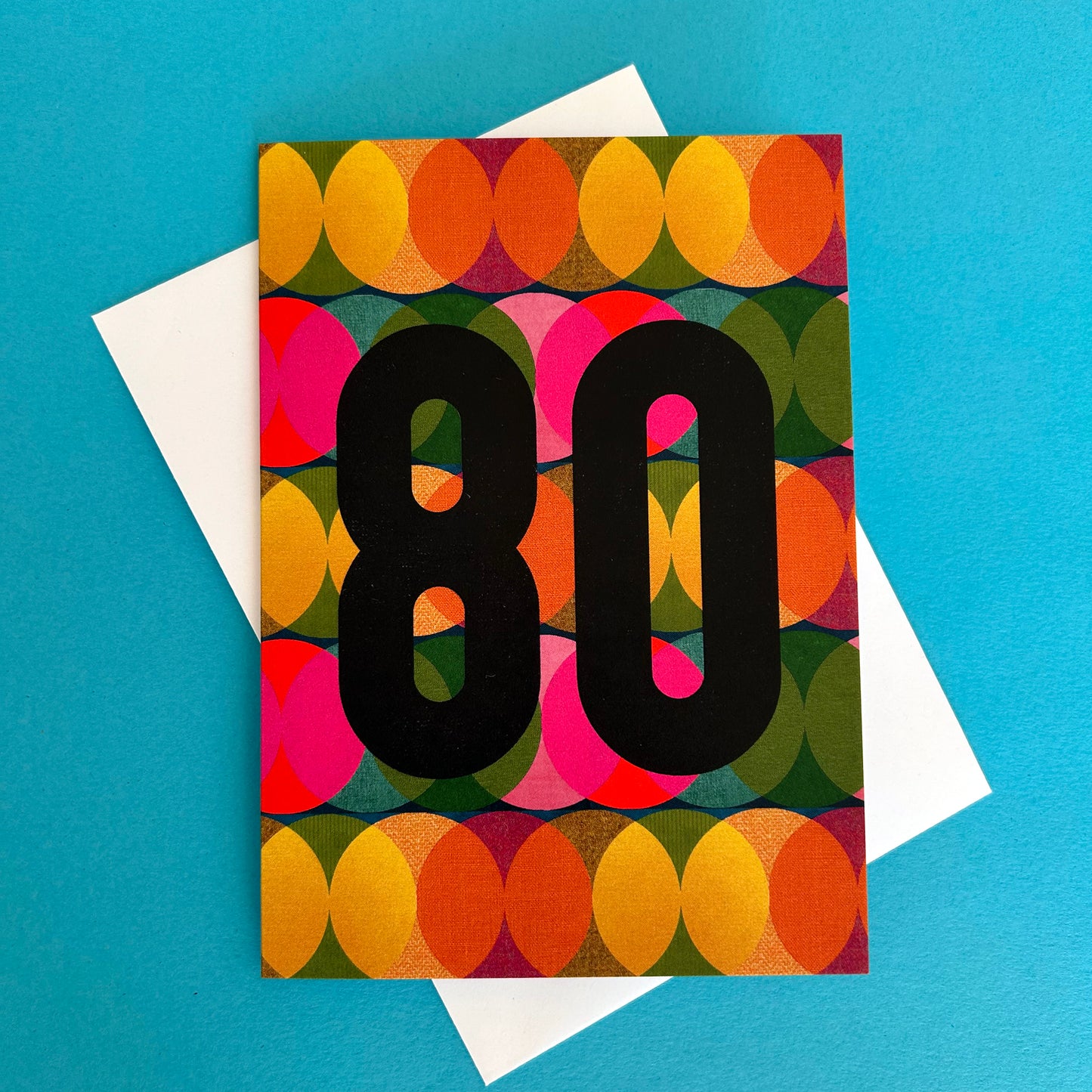 A bright greeting card with a colourful retro geometric pattern of overlapping circles in orange, yellow, pink, green, and purple, featuring the number '80' in bold black text. The card is displayed on top of a white envelope against a solid bright blue background.