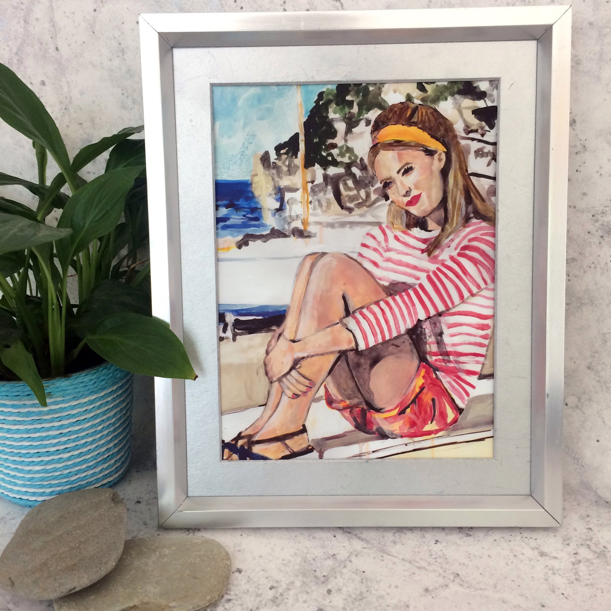 Image shows a framed illustration of a woman with a red and white striped top, red shorts and a yellow headband  sitting on a wall by the coast with rocks and a blue sea in the background.