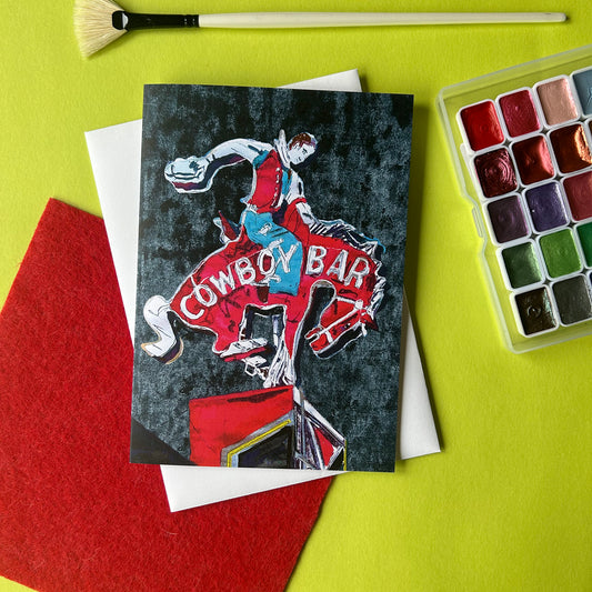 Card with cowboy design and 'Cowboy Bar' text on a green surface with art supplies.