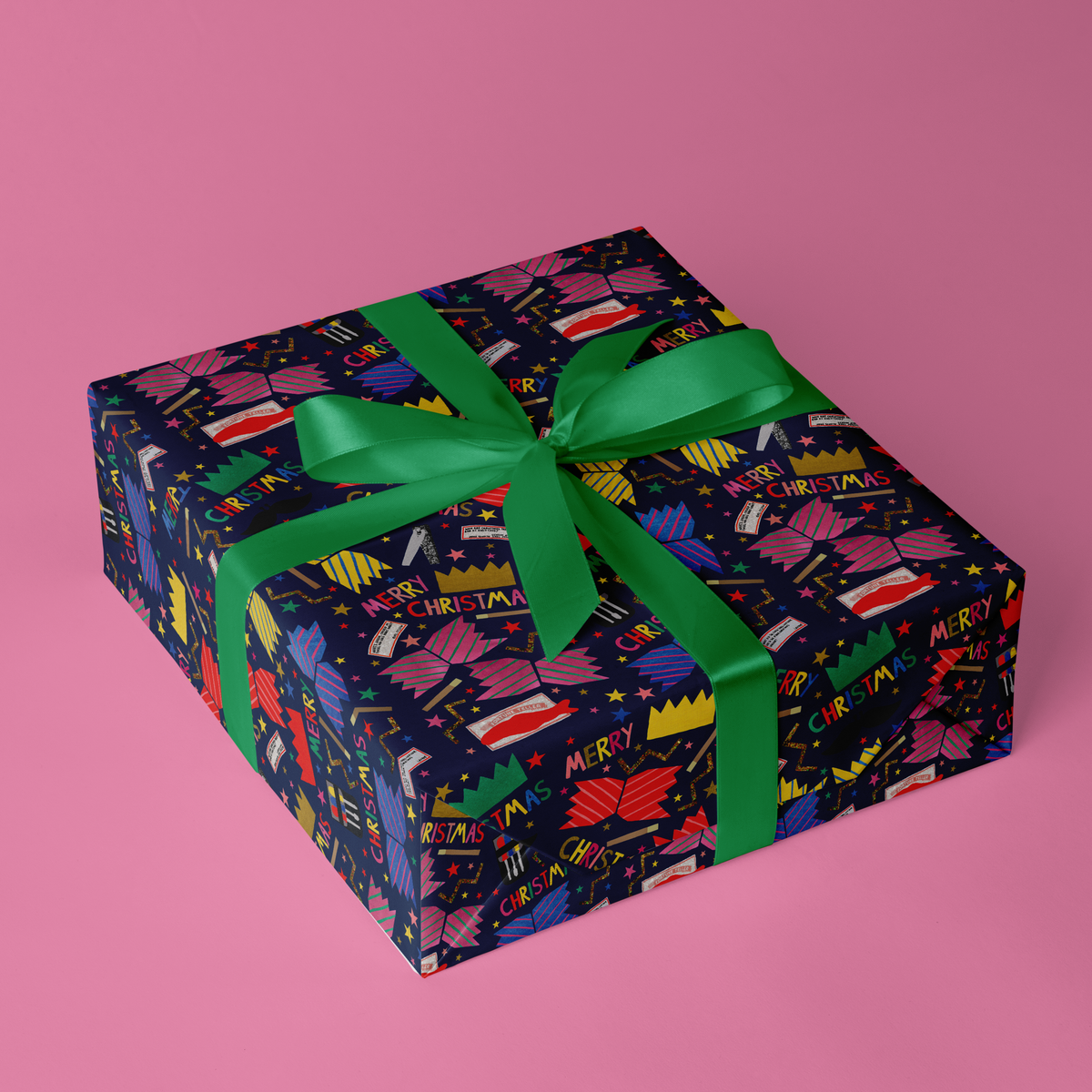 A square gift box wrapped in dark navy Christmas-themed wrapping paper with a colourful festive cracker pattern and tied with a bright green satin ribbon, set against a solid pink background.