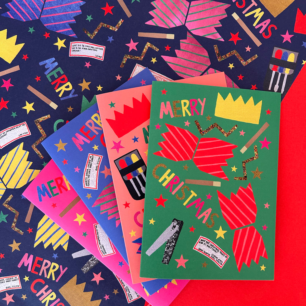 A set of colourful Christmas greeting cards arranged in a fan-like stack on top of patterned wrapping paper. The front card is green with red crackers, gold stars, and the words 'MERRY CHRISTMAS' and a yellow crown.