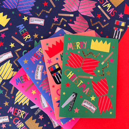 A set of colourful Christmas greeting cards arranged in a fan-like stack on top of patterned wrapping paper. The front card is green with red crackers, gold stars, and the words 'MERRY CHRISTMAS' and a yellow crown.