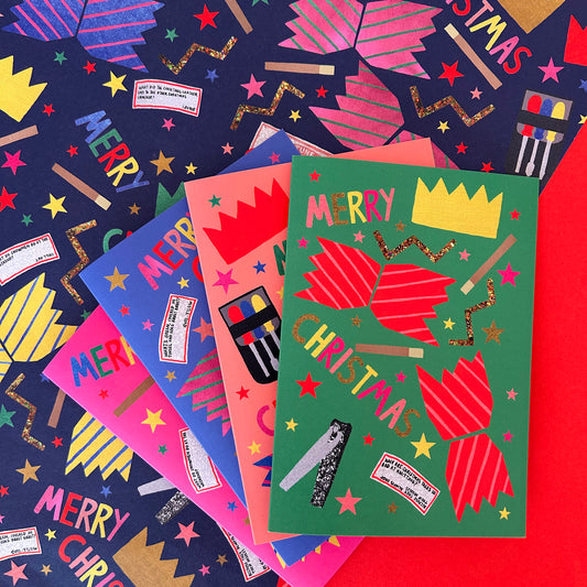 A set of colourful Christmas greeting cards arranged in a fan-like stack on top of patterned wrapping paper. The front card is green with red crackers, gold stars, and the words 'MERRY CHRISTMAS' and a yellow crown.