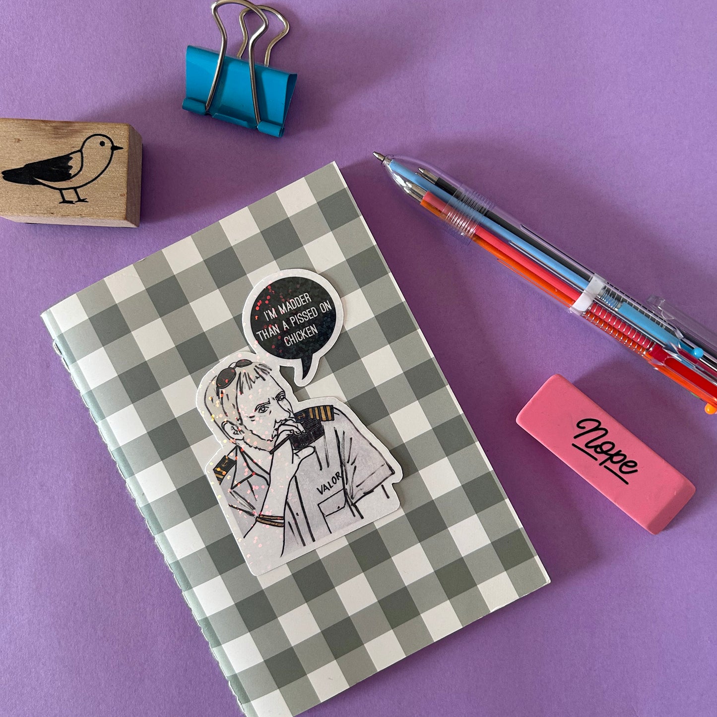 Checkered notebook with a Captain Lee vinyl holographic sticker and text bubble, surrounded by stationery items on a purple background.