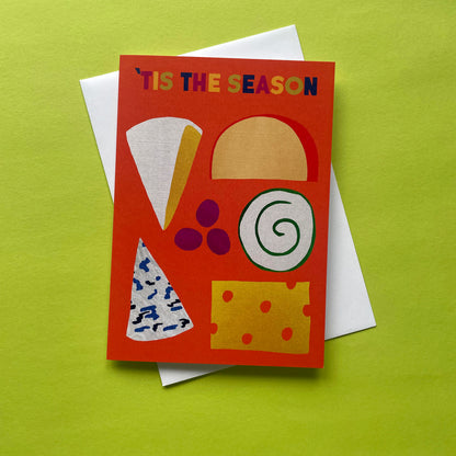 An orange greeting card with the text "IT'S THE SEASON" and colourful cheese-themed illustrations, layered over a white envelope on a bright yellow-green background.