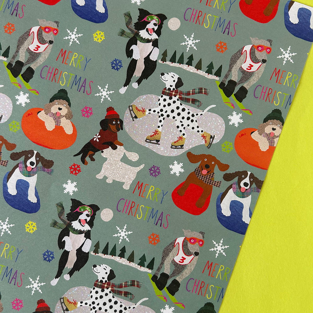 A close-up of a Christmas-themed dog print fabric or wrapping paper featuring illustrated dogs in winter activities on a muted sage green background.