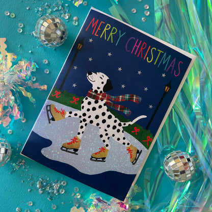 A festive Christmas greeting card featuring a Dalmatian dog ice skating at night under a rainbow 'MERRY CHRISTMAS' header. The card is styled on a bright teal background with disco balls, tinsel, and sequins.