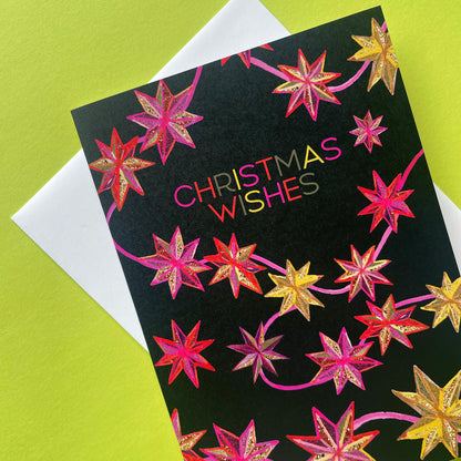 A Christmas greeting card with a black background and colourful metallic star designs, shown with a white envelope on a bright yellow-green surface. The card reads 'CHRISTMAS WISHES' in multicoloured lettering.
