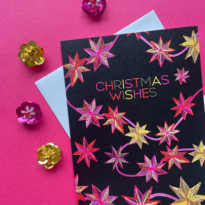 A Christmas greeting card with a black background and colourful metallic star illustrations, reading 'CHRISTMAS WISHES' in multicoloured lettering. The card is styled on a bright pink background with an envelope and decorative metallic flower props.