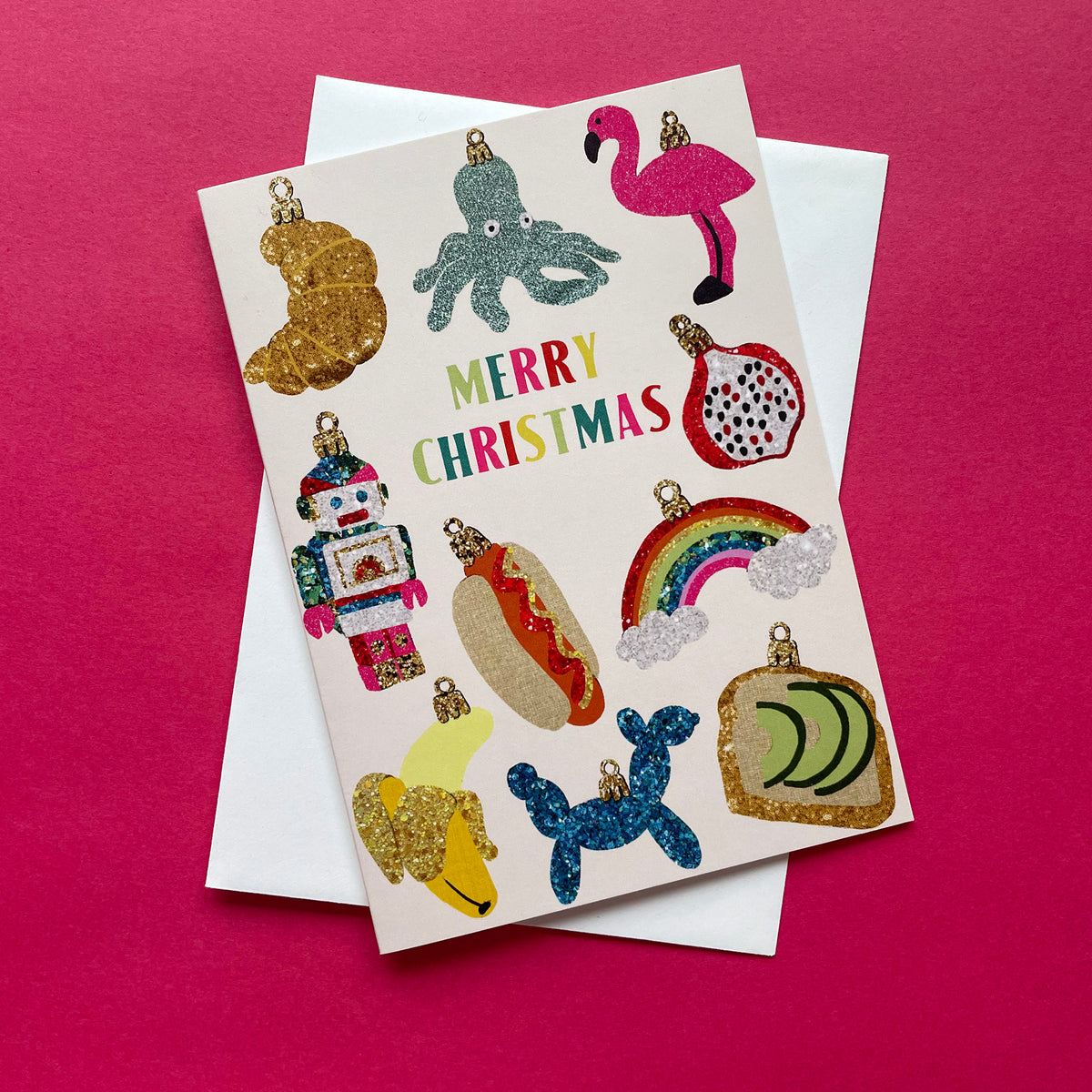 A Christmas greeting card featuring colourful glitter-style holiday ornaments and the words “MERRY CHRISTMAS” in multicoloured lettering, placed on top of a white envelope against a bright pink background.