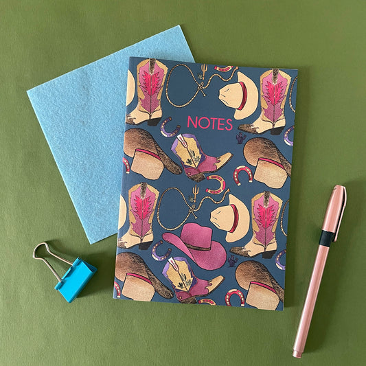 Image of a blue notebook with a western inspired pattern of illustrated cowboy boots, hats, horseshoes and lassos with the word Notes in pink writing.
