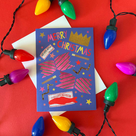 A festive Christmas greeting card with a blue background, pink striped cracker, and 'MERRY CHRISTMAS' in colourful letters, styled on a white envelope against a red background with Christmas lights.