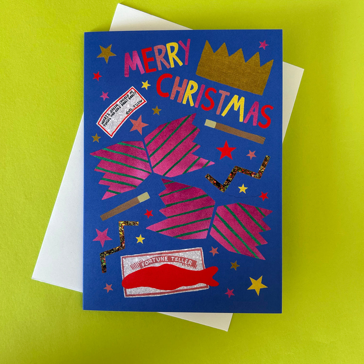 A Christmas greeting card with a blue background and colourful festive illustrations, placed on top of a white envelope against a bright yellow-green surface. The card reads 'MERRY CHRISTMAS' with stars, christmas cracker, a gold crown, and a joke.
