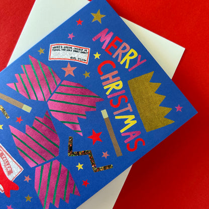 A blue Christmas greeting card with colourful lettering reading 'MERRY CHRISTMAS,' pink striped cracker, gold stars, and a gold crown, photographed on a red background with part of a white envelope visible.