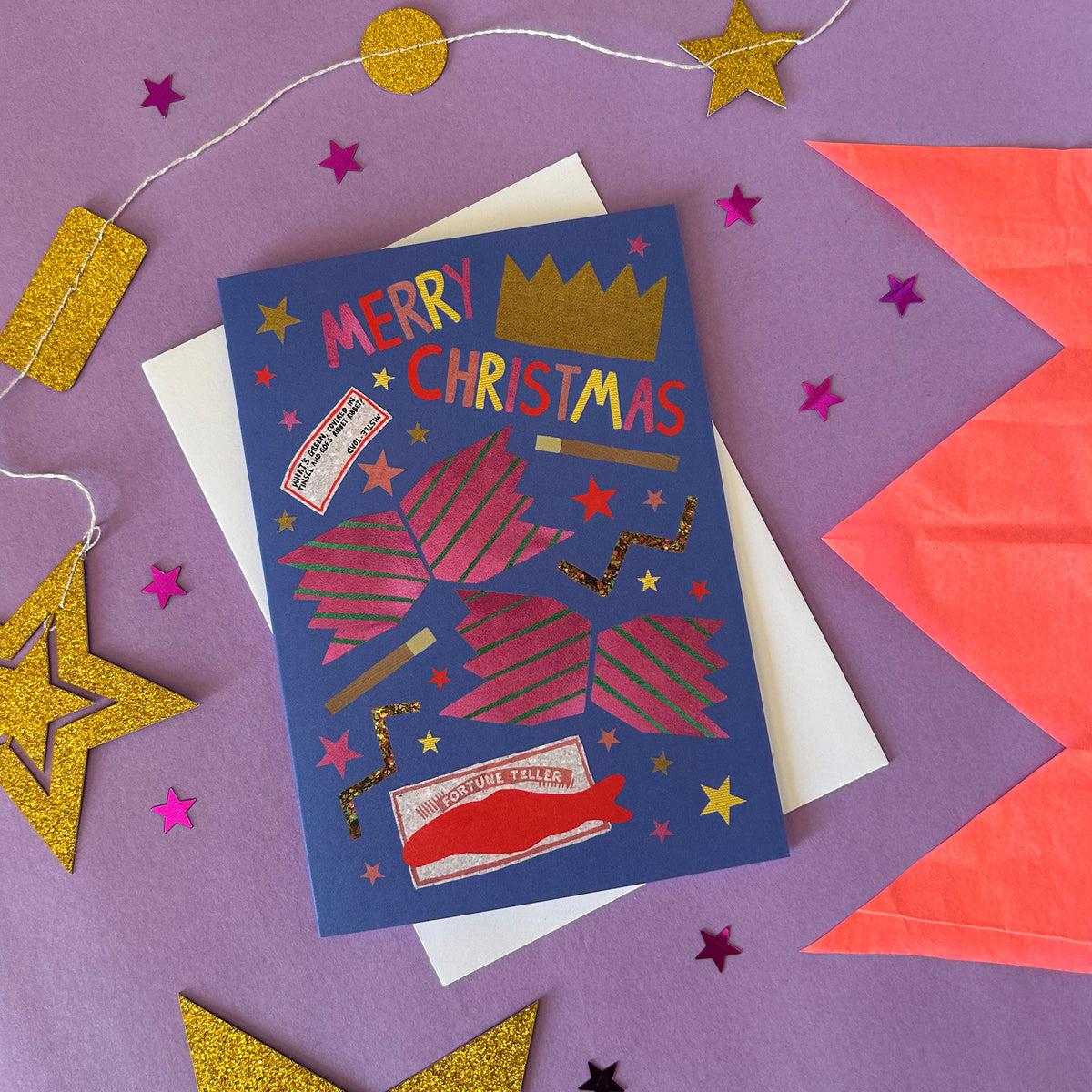 A blue Christmas card with pink cracker design and fun details, displayed on top of a white envelope against a purple background decorated with gold and pink paper stars.