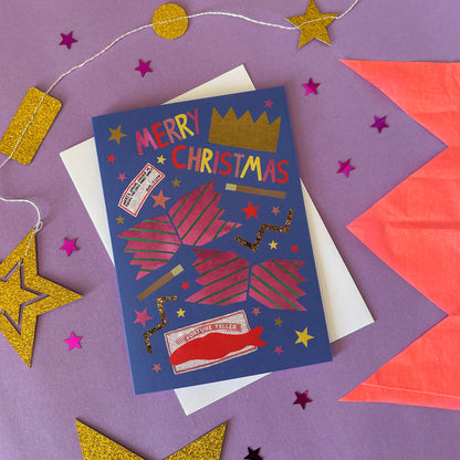A blue Christmas card with pink cracker design and fun details, displayed on top of a white envelope against a purple background decorated with gold and pink paper stars.