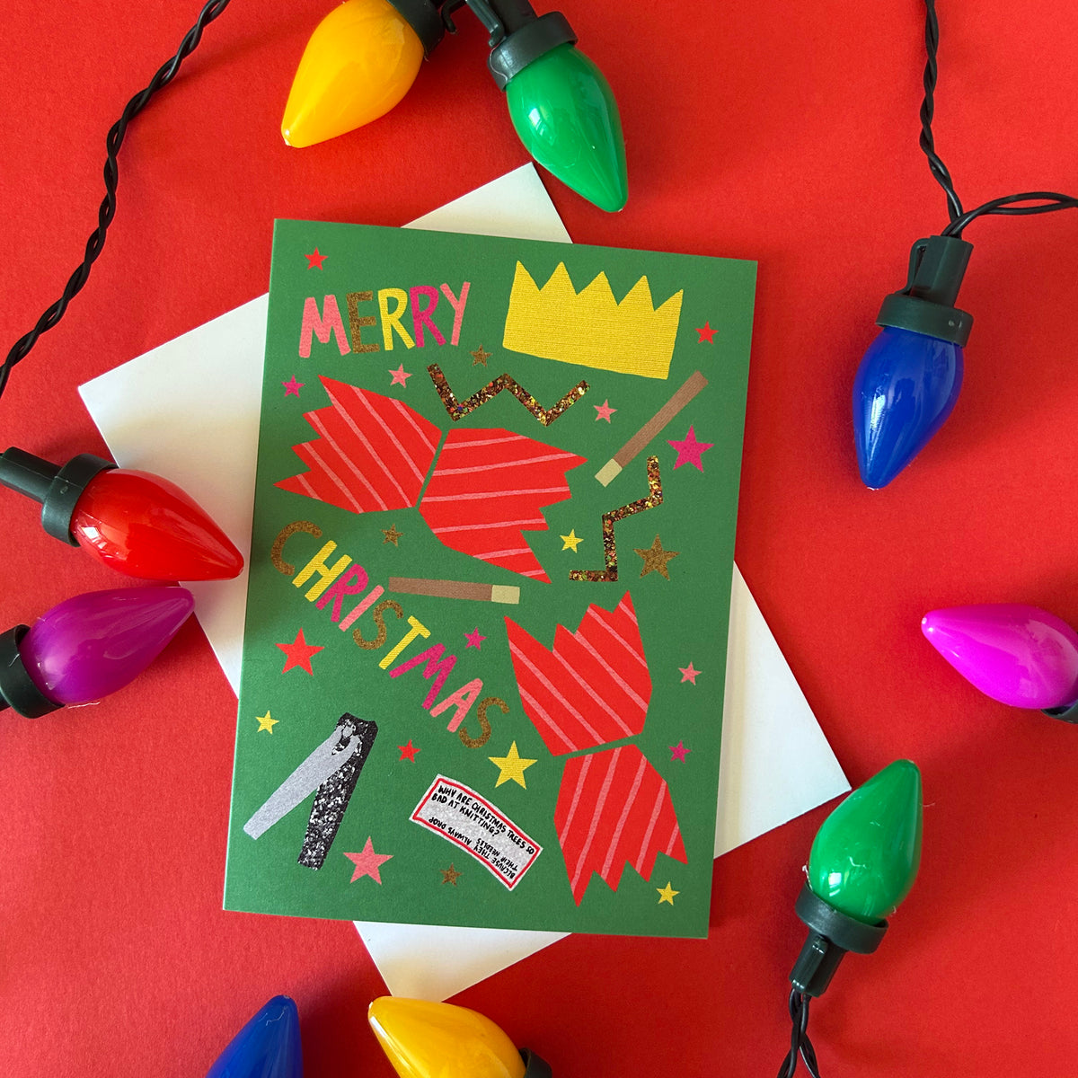A green Christmas greeting card with festive red, gold, and pink cracker contents styled on a red background with white envelopes and colourful Christmas lights.
