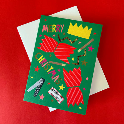 A green Christmas greeting card with a colourful festive cracker design, including the words 'MERRY CHRISTMAS,' a yellow crown, stars, and glittery details, shown on top of a white envelope against a red background.