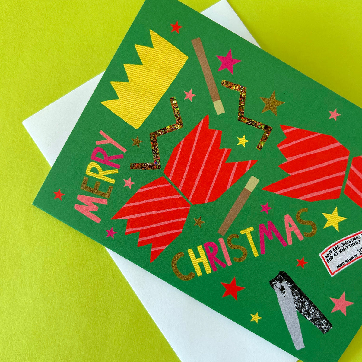 A festive Christmas card featuring colourful illustrations of crackers, stars, and the words 'MERRY CHRISTMAS' on a green background, photographed on a bright lime surface with a white envelope partially visible.