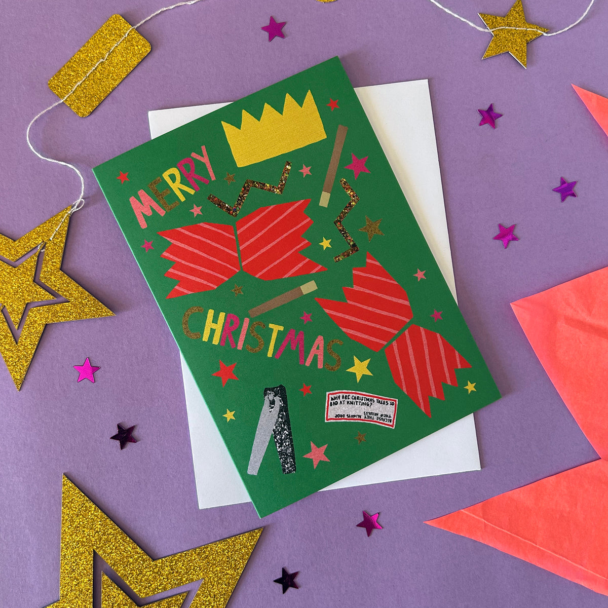 A festive Christmas card with a green background, red striped cracker illustrations, and gold glitter accents, styled on a purple surface with gold and pink star decorations.