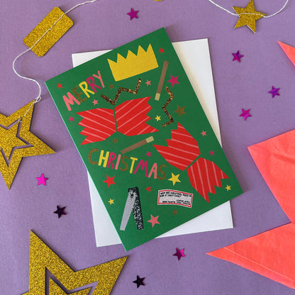 A festive Christmas card with a green background, red striped cracker illustrations, and gold glitter accents, styled on a purple surface with gold and pink star decorations.