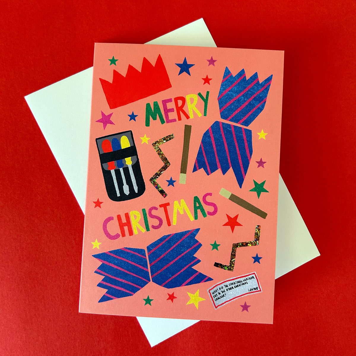 A Christmas greeting card with a peach background and colourful festive illustrations, shown with a white envelope on a red surface. The card reads 'MERRY CHRISTMAS' and features blue striped cracker and small multicoloured stars.