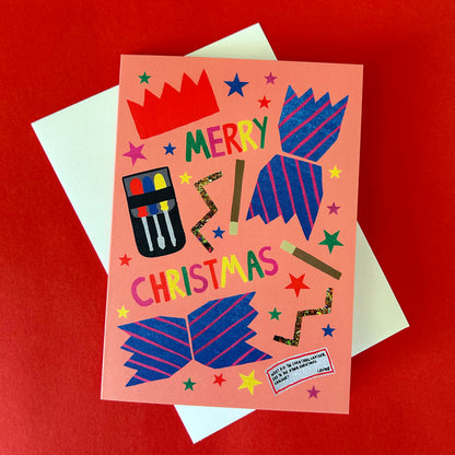 A Christmas greeting card with a peach background and colourful festive illustrations, shown with a white envelope on a red surface. The card reads 'MERRY CHRISTMAS' and features blue striped cracker and small multicoloured stars.
