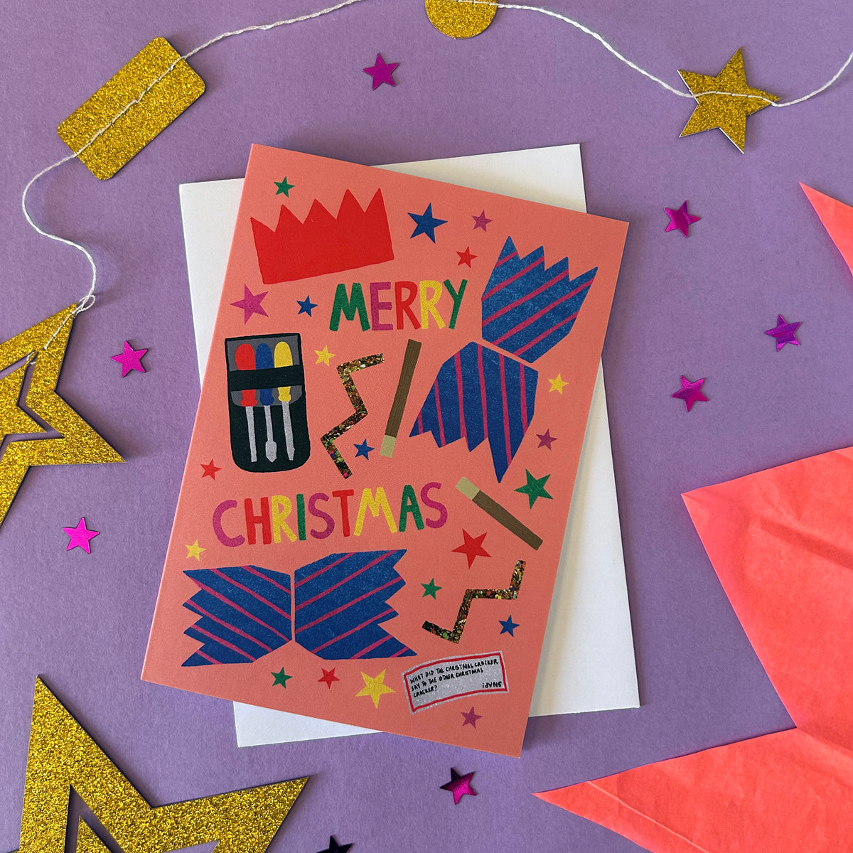 A festive Christmas greeting card with a coral-pink background and colourful illustrations of crackers, stars, and the words 'MERRY CHRISTMAS', styled on a purple surface with gold glitter star garlands and orange paper accents.