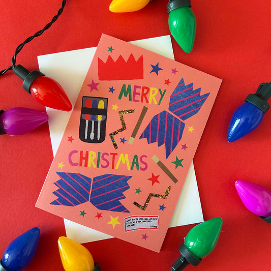 A festive Christmas card with a peach background and colourful cracker illustrations, styled on a red surface with multicoloured Christmas lights and a white envelope.