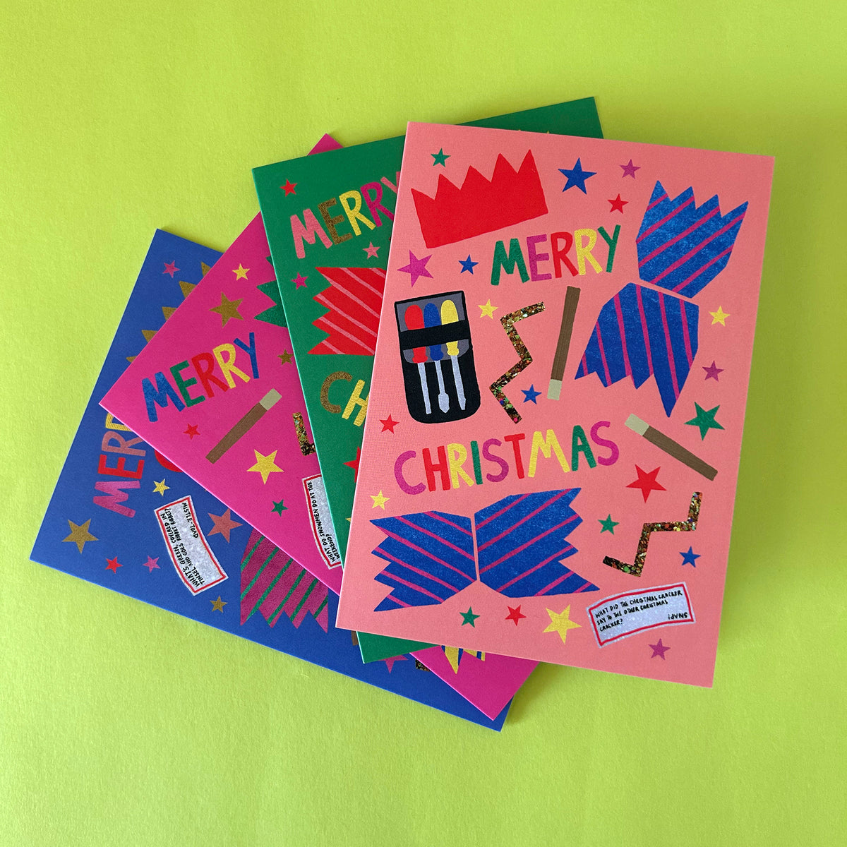 A stack of colourful Christmas greeting cards fanned out on a bright lime green background. The top card is pink with a red crown, blue cracker, and multicoloured stars reading 'MERRY CHRISTMAS'; beneath it are blue, pink, and green cards with similar festive designs.