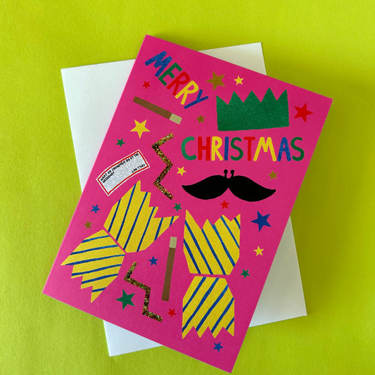 A bright pink Christmas greeting card with colorful cutout-style graphics and the words 'MERRY CHRISTMAS' on the front, layered over a white envelope against a lime green background.