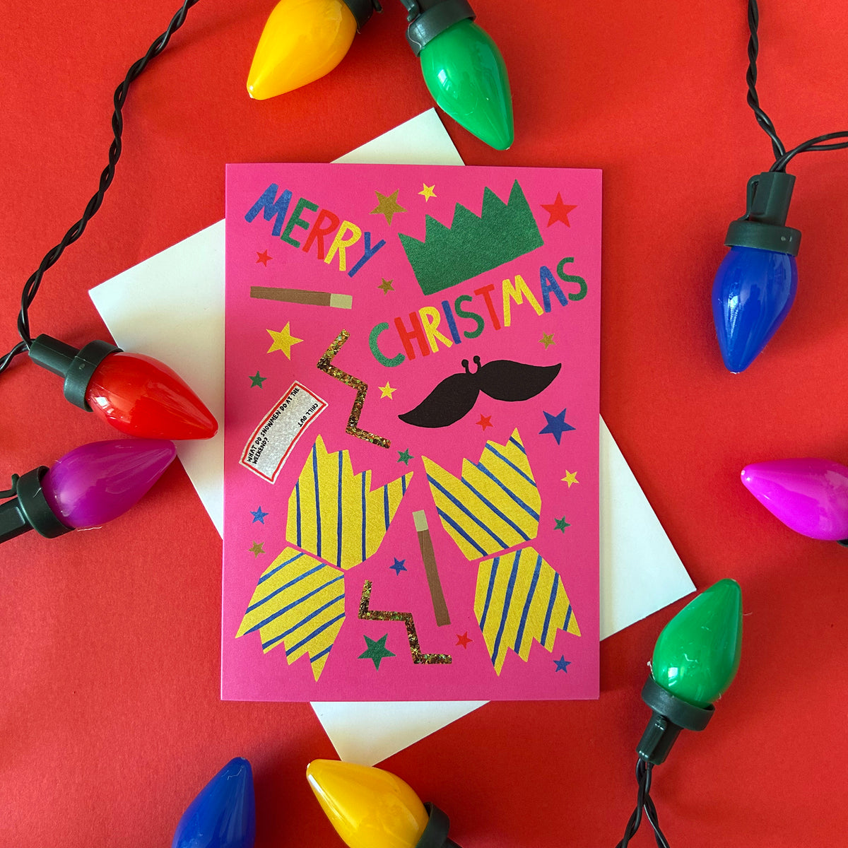 A pink Christmas greeting card with colourful cracker design styled on a red background with white envelopes and festive multicolour Christmas lights.