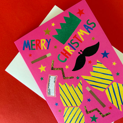 A handmade pink Christmas card with colourful cracker decorations, featuring the text 'MERRY CHRISTMAS' and a black moustache, layered over a white envelope on a red background.