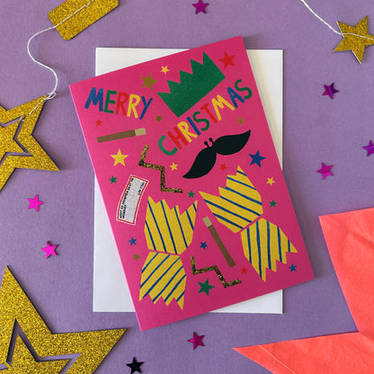 A festive Christmas greeting card with a bright pink background and colourful cracker illustrations, placed on top of a white envelope against a purple surface with glittery star decorations.