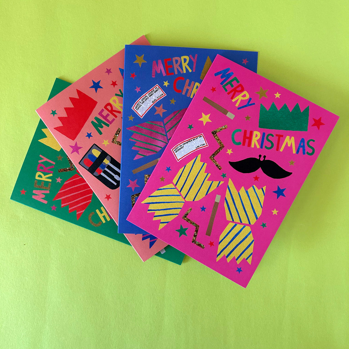 Four colourful Christmas greeting cards fanned out on a bright lime green background. The cards feature festive cracker designs including stars, stripes, a moustache, and paper hat in pink, blue, green, and coral colours.