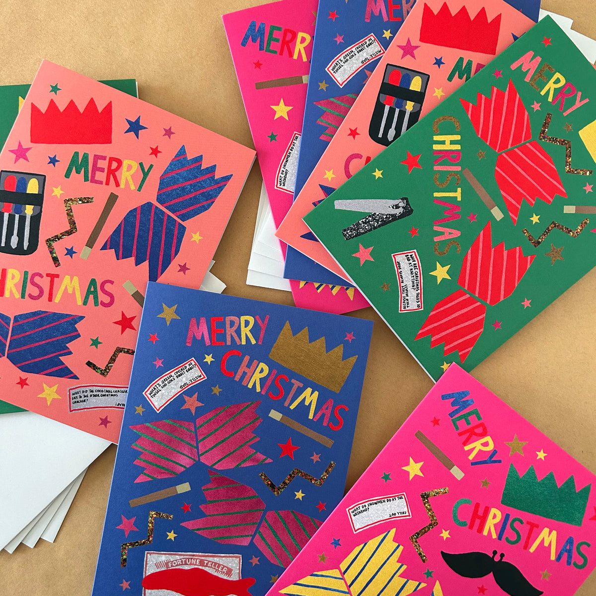 A collection of colourful handmade Christmas cards featuring crackers, stars, crowns, and the words 'MERRY CHRISTMAS' in various bright colours on a tan background.