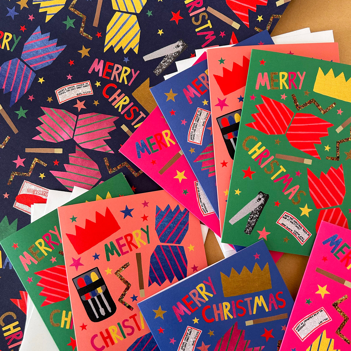 A pile of colourful Christmas cracker themed greeting cards and wrapping paper on a wooden surface. The designs feature bright cracker illustrations, gold glitter crowns, and festive text 'MERRY CHRISTMAS'
