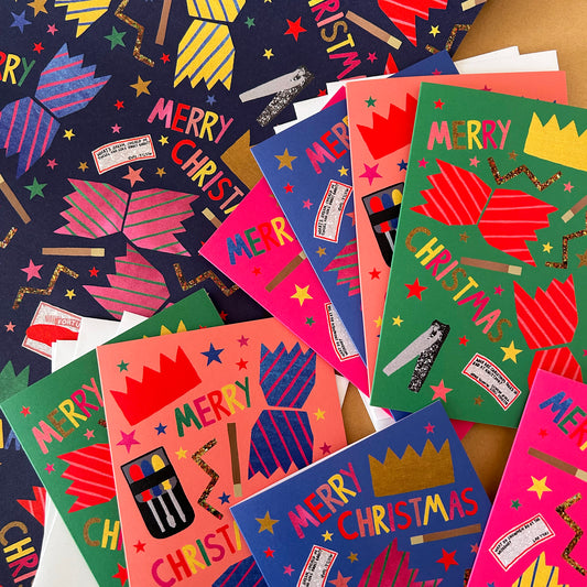 A pile of colourful Christmas cracker themed greeting cards and wrapping paper on a wooden surface. The designs feature bright cracker illustrations, gold glitter crowns, and festive text 'MERRY CHRISTMAS'
