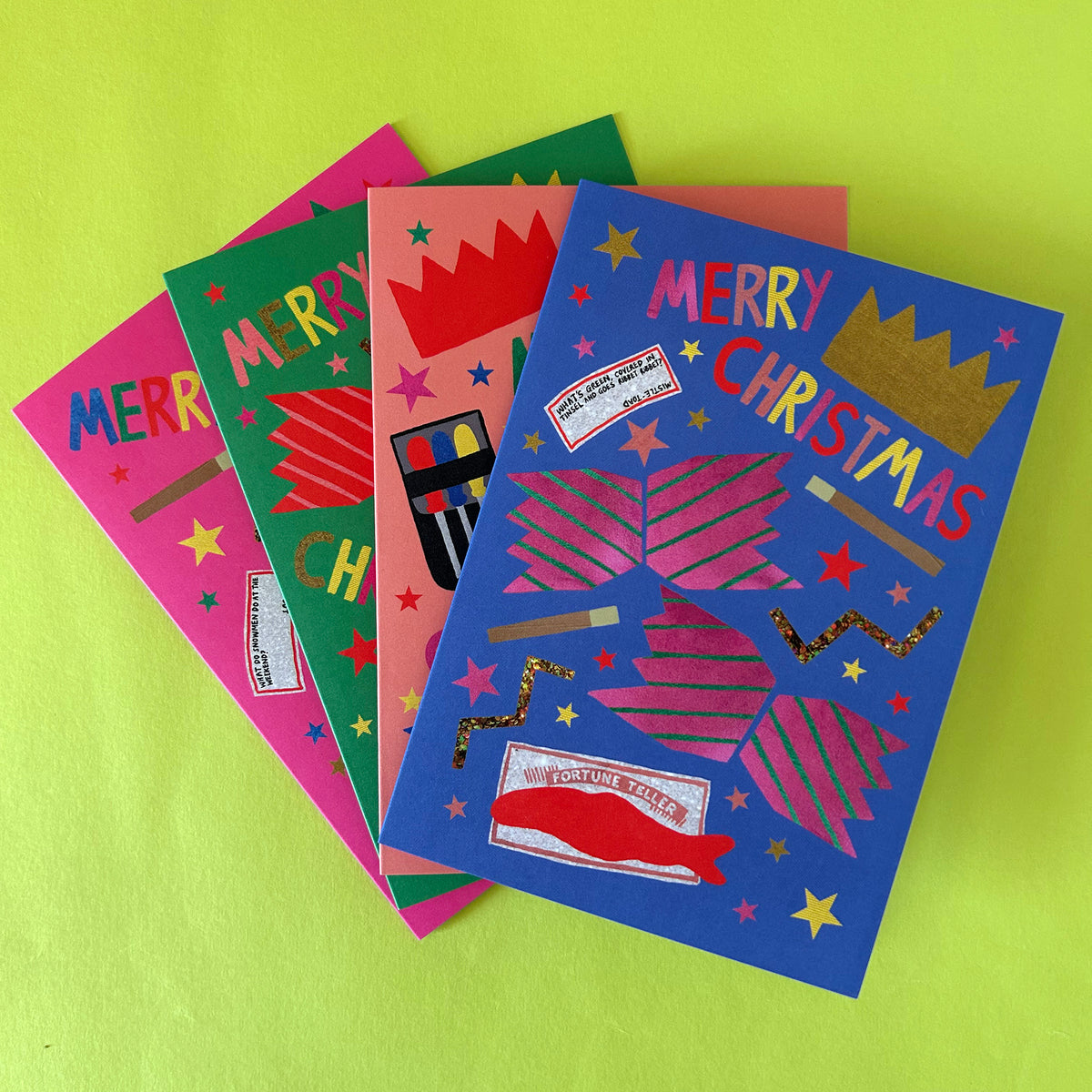 Four colourful Christmas greeting cards fanned out on a bright lime green background. The cards feature festive designs with 'MERRY CHRISTMAS'  crackers, prizes, and gold foil accents in pink, green, coral, and blue.
