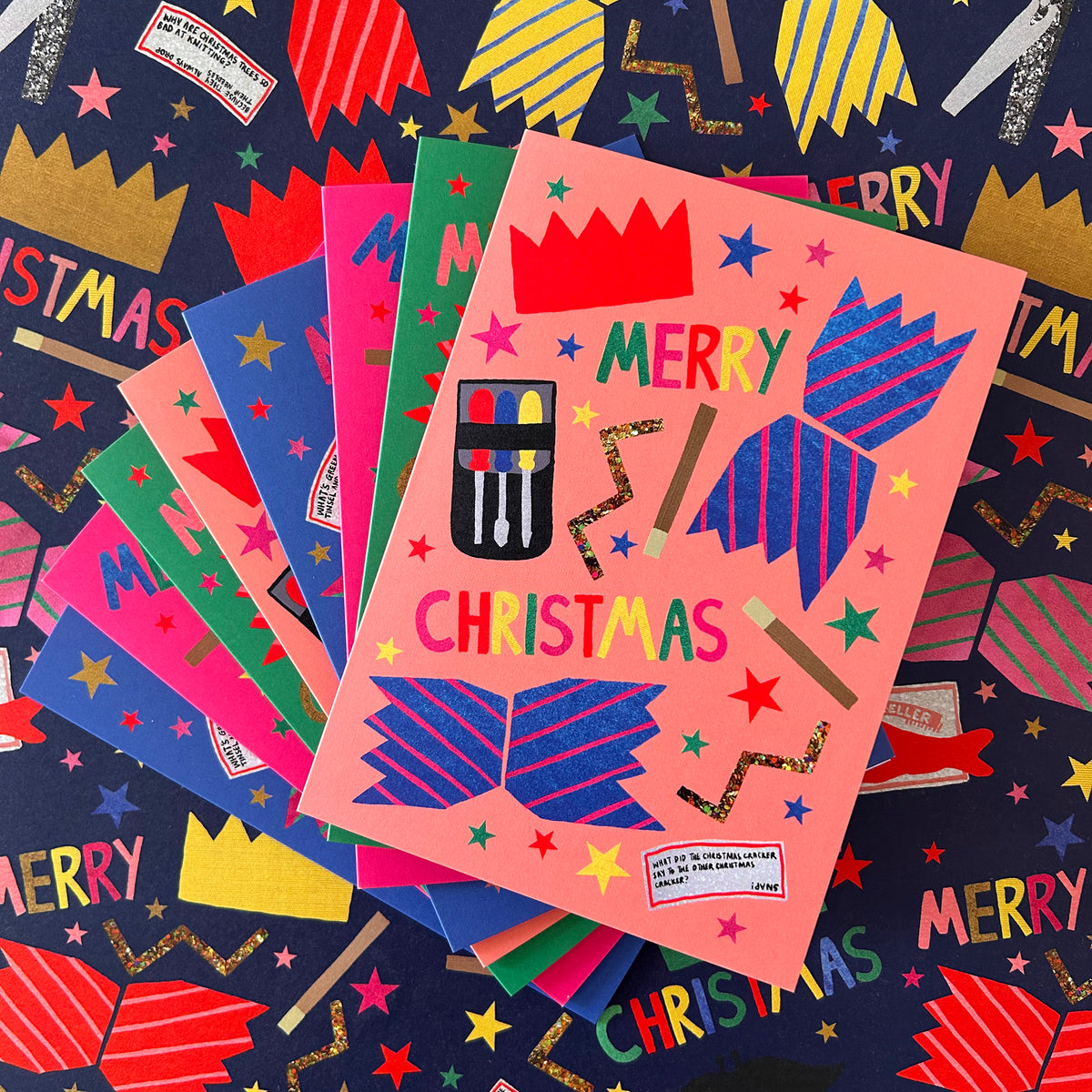 A stack of colourful Christmas greeting cards fanned out on a dark navy matching gift wrap with festive illustrations. The top card is peach-pink with blue striped crackers, gold glitter stars, and the words 'MERRY CHRISTMAS' in multicoloured letters.