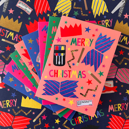 A stack of colourful Christmas greeting cards fanned out on a dark navy matching gift wrap with festive illustrations. The top card is peach-pink with blue striped crackers, gold glitter stars, and the words 'MERRY CHRISTMAS' in multicoloured letters.