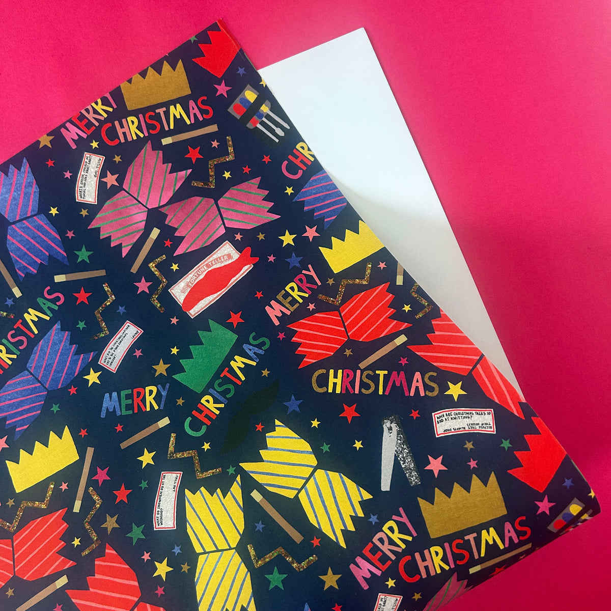 A close-up of a Christmas-themed wrapping paper with a dark navy background and colourful festive illustrations, including 'MERRY CHRISTMAS' and brightly coloured crackers on a bright pink background.