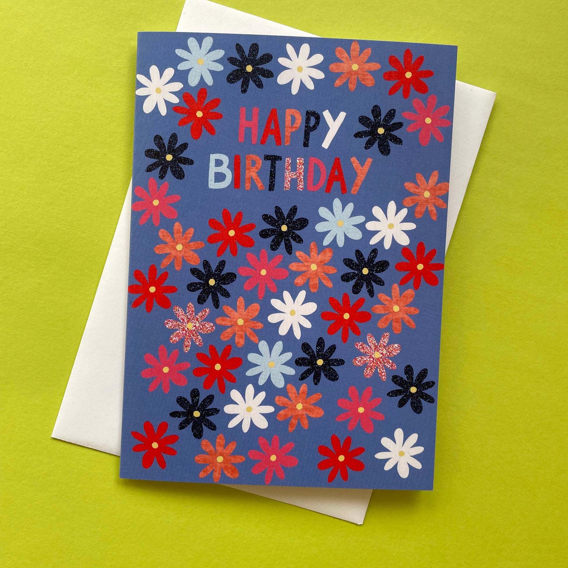 Colourful birthday card with flowers and 'Happy Birthday' text on a blue background, placed on a green surface.